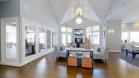 The Submit Clubhouse is a thoughtfully designed community gathering space that combines elegance with comfort. The main lounge immediately impresses with its dramatic vaulted ceilings that soar to a sharp peak, creating an airy, open atmosphere. A striking geometric chandelier hangs at the center, complemented by a circular skylight window that floods the room with natural light. Large, multi-pane windows line the walls on multiple sides, further brightening the interior and offering views of the surrounding outdoors. Rich hardwood flooring runs throughout, adding warmth and character to the space. The seating area is arranged around a central console table, featuring plush gray sofas and vibrant orange leather ottomans that introduce a pop of color and an inviting, modern aesthetic. A wall-mounted flat-screen television above a fireplace serves as a focal point for the gathering area. Adjacent to the main lounge, glass-paneled doors reveal a fitness area equipped with exercise machines, seamlessly integrating wellness amenities into the clubhouse experience. The overall ambiance is bright, sophisticated, and welcoming — an ideal setting for both relaxed socializing and community events.