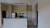 This single-floor space centers around an open-concept kitchen that serves as the heart of the layout. The kitchen is well-equipped with stainless steel appliances, including a refrigerator, a built-in microwave, and a range, all seamlessly integrated into the design. Light wood cabinetry lines the upper walls, providing generous storage space while contributing a warm, neutral aesthetic to the room.

A peninsula-style breakfast bar extends outward, creating a natural boundary between the kitchen and the adjacent living area, while also offering a convenient spot for casual dining or meal prep. The countertops are finished in a clean, light tone that complements the overall color palette.

Recessed lighting and a track light fixture illuminate the kitchen area effectively, ensuring a bright and functional workspace. The flooring transitions between the kitchen and the adjoining hallway, adding subtle visual definition to the space. A hallway visible from the main area leads to additional rooms, suggesting a practical and straightforward floor plan. The neutral wall tones throughout create a cohesive and understated ambiance suitable for a variety of interior styles.