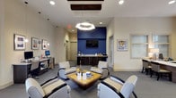 Lobby 2 is a thoughtfully designed lounge space that combines contemporary style with practical amenities. The room is anchored by a striking cobalt blue accent wall, which serves as a bold focal point and provides a vibrant contrast to the neutral cream and gray tones throughout the space. A wall-mounted flat-screen television is positioned on this accent wall, adding a functional entertainment element to the area.

The seating arrangement centers around a rustic wooden coffee table, surrounded by a set of modern, light gray armchairs with sleek black accents, creating an inviting and cohesive conversation area. Overhead, a distinctive geometric pendant light fixture illuminates the space, complemented by recessed lighting throughout the ceiling. A coffered ceiling detail with rich dark wood trim adds an architectural element of depth and character.

Along one wall, a service counter with dark cabinetry and a light stone countertop offers a refreshment or service station, enhancing the lobby's utility. A workstation with a computer terminal is also available nearby. Framed artwork adorns the walls, contributing to the polished, welcoming atmosphere of this well-appointed lobby space.