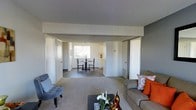 This single-floor residence is designed with an open, flowing layout that connects the main living areas seamlessly. The living room serves as the central hub of the home, featuring plush carpeting in warm neutral tones, a textured ceiling, and ample space for comfortable furnishings. Large sliding closet doors provide generous storage directly off the living area, adding to the home's practicality.

The space opens naturally into a combined dining area, where a compact dining set is positioned near a window that allows natural light to filter into the room. This creates a bright, welcoming atmosphere in the dining space. Just beyond, the kitchen is visible with white cabinetry that contributes to a clean, classic aesthetic.

The layout benefits from multiple doorways and corridors that suggest well-defined private areas, likely including bedrooms and bathrooms, accessible from the main living space. The consistent use of light gray walls and neutral carpeting throughout creates a cohesive, understated palette that allows for versatile interior styling.

Overall, this residence offers a practical and comfortable floor plan with an efficient use of space, good natural light in the dining and kitchen areas, and a neutral design foundation that is both functional and adaptable.