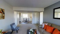 This single-floor residence is designed with an open, flowing layout that connects the main living areas seamlessly. The living room serves as the central hub of the home, featuring plush carpeting in warm neutral tones, a textured ceiling, and ample space for comfortable furnishings. Large sliding closet doors provide generous storage directly off the living area, adding to the home's practicality.

The space opens naturally into a combined dining area, where a compact dining set is positioned near a window that allows natural light to filter into the room. This creates a bright, welcoming atmosphere in the dining space. Just beyond, the kitchen is visible with white cabinetry that contributes to a clean, classic aesthetic.

The layout benefits from multiple doorways and corridors that suggest well-defined private areas, likely including bedrooms and bathrooms, accessible from the main living space. The consistent use of light gray walls and neutral carpeting throughout creates a cohesive, understated palette that allows for versatile interior styling.

Overall, this residence offers a practical and comfortable floor plan with an efficient use of space, good natural light in the dining and kitchen areas, and a neutral design foundation that is both functional and adaptable.