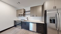 This thoughtfully designed single-floor space showcases a clean, modern aesthetic with high-quality finishes throughout. The open-concept kitchen is a standout feature, boasting two-tone cabinetry with dark lower cabinets and warm taupe upper cabinets accented by matte black hardware. A white subway tile backsplash, light quartz countertops, and a full suite of stainless steel appliances — including a range, built-in microwave, dishwasher, and French door refrigerator — complete the functional and stylish cooking space. Open shelving adds a contemporary touch alongside ample cabinet storage. Light-toned luxury vinyl plank flooring flows seamlessly through the main living areas, creating a cohesive and airy feel. The bathroom features large-format gray tile flooring, a generously sized light wood vanity with quartz countertops, matte black hardware, a wide framed mirror, and bright vanity lighting. A bold dark-paneled door adds a striking design contrast. The living area is bright and open, highlighted by recessed lighting, a modern ceiling fan, and sliding glass doors that lead to a private balcony, inviting natural light and outdoor views into the space. Clean white walls and dark baseboards tie the overall design together with a polished, contemporary look.