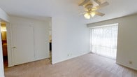 This single-story space presents a clean, functional layout designed for comfortable everyday living. The main living area is finished with wall-to-wall beige carpet and painted in crisp white, creating a bright and neutral atmosphere. A ceiling fan with an integrated light fixture is centrally mounted, providing both ventilation and ambient lighting. A large window dressed with vertical blinds allows generous natural light to fill the room while offering privacy control. The white walls and light-toned carpet contribute to an open, airy feel throughout. An open passageway connects the living area to an adjacent kitchen, which is partially visible and features cabinetry and appliances. The textured ceiling adds a subtle architectural detail consistent throughout the space. The overall layout is straightforward and practical, with a simple, understated aesthetic that offers a versatile canvas for personalization.