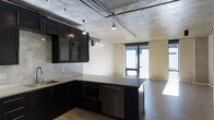 This single-floor space makes a striking impression with its industrial-modern aesthetic, defined by a raw exposed concrete ceiling that serves as a bold architectural centerpiece. Suspended pendant lights and visible conduit runs add to the urban loft character, lending an authentic industrial edge throughout.

The open-plan kitchen is a standout feature, outfitted with dark matte cabinetry that extends to the upper walls and is complemented by glass-fronted upper cabinets for a refined touch. Light-toned countertops provide a crisp contrast against the dark finishes, while a marble-effect tile backsplash adds texture and visual interest. A kitchen island with matching dark cabinetry and a dark countertop anchors the space and provides additional workspace. Stainless steel appliances, including a built-in oven and dishwasher, complete the kitchen's functional and contemporary design.

A wall-mounted split-system air conditioning unit ensures year-round comfort. The living area flows seamlessly from the kitchen, bathed in abundant natural light streaming through large floor-to-ceiling windows that frame bright outdoor views. Warm wood-toned flooring runs throughout, softening the industrial elements and tying the space together cohesively. The overall ambiance is sleek, urban, and inviting.