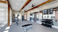 Located at 11 Parklands Drive in Bluffton, South Carolina, this single-story fitness center offers a comprehensive workout facility designed with both functionality and aesthetic appeal in mind. The space is distinguished by its striking exposed dark wood beam ceilings, which lend a warm, rustic character to the otherwise bright and modern interior. Ceiling fans are positioned throughout, ensuring comfortable airflow during workouts.

The main cardio area is generously sized, housing multiple treadmills, elliptical trainers, and stair climbers arranged to maximize space and usability. Large windows line the walls, flooding the room with abundant natural light and offering views of the surrounding landscape. A dedicated cycling studio features several spin bikes and a rack of kettlebells, providing ample space for group or individual cycle training, with hardwood flooring adding a polished finish to that section.

The strength training zone includes a multi-station cable machine, adjustable weight benches, and a full rack of dumbbells, catering to a wide range of resistance training needs. Wall-mounted mirrors enhance the sense of space and allow for proper form checks. Stability balls and additional fitness accessories are also available. The overall ambiance is clean, bright, and inviting, making this fitness center a well-rounded facility for residents of the Parklands community.