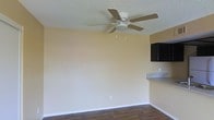 This single-floor space offers a straightforward, functional layout with a clean and practical design. The main living area is defined by warm beige walls and dark wood-style laminate flooring, which together create a grounded, neutral aesthetic. A five-blade ceiling fan with an integrated light fixture is centrally mounted on a textured popcorn ceiling, providing both air circulation and ambient lighting to the room.

Adjacent to the living area, a partial view of the kitchen reveals dark upper cabinetry with a sleek finish, paired with a granite-style countertop that extends as a serving or workspace ledge. This transitional counter element visually connects the kitchen to the living space, lending a sense of openness to the overall floor plan.

White baseboards provide a crisp contrast against both the flooring and the walls, contributing to a tidy, well-defined finish throughout the space. Electrical outlets are positioned at standard intervals along the walls, reflecting practical, everyday usability. The overall ambiance is modest and move-in ready, suited for straightforward, comfortable living.