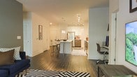 This single-floor space showcases a thoughtfully designed open-concept layout that connects the living, dining, and kitchen areas into one cohesive environment. Rich, dark hardwood flooring runs throughout, providing a warm contrast to the bright white walls and cabinetry that define the interior aesthetic.

The kitchen is well-appointed with stainless steel appliances, including a full-size refrigerator and dishwasher, complemented by white upper cabinets and wood-toned lower cabinetry. A central island with bar-height seating serves as both a prep area and casual dining space, illuminated by pendant lighting above and a track lighting system that ensures ample brightness across the kitchen zone.

Adjacent to the kitchen, a dedicated workspace area features a desk and ergonomic seating, making it a functional corner for productivity. Bold black-and-white geometric area rugs anchor the living and kitchen zones, adding visual interest to the open floor plan.

The layout benefits from clean sightlines across all areas, creating a sense of spaciousness throughout. White walls and recessed lighting further enhance the bright, airy ambiance that characterizes this well-organized, modern space.