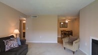 This single-story space features an open-concept layout that seamlessly connects the living room and dining area, creating a cohesive and functional flow throughout. The living area is finished with plush carpeting in a neutral tone, providing a comfortable and inviting atmosphere. Warm beige walls and white baseboards give the space a clean, understated aesthetic.

A ceiling fan with a light fixture serves the dining area, which accommodates a wooden table and chair set. A wall-mounted mirror adds depth to the dining space, while hardwood-style flooring grounds the area and contrasts nicely with the carpeted living room. Overhead HVAC venting is integrated into the ceiling, ensuring practical climate control throughout.

A hallway visible from the living area leads to additional rooms, with glimpses of what appears to be a kitchen with cabinetry in warm wood tones. The textured ceiling adds a subtle design element consistent throughout the space. Overall, the layout is straightforward and functional, with neutral finishes that provide a versatile backdrop suitable for a variety of interior styles.