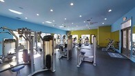 This fitness center is a thoughtfully designed, single-floor facility built to support a comprehensive workout experience. The space is immediately welcoming, featuring a vibrant color palette with bold teal and lime green accent walls that energize the environment and give it a modern, contemporary feel.

The layout is open and spacious, with ample room between equipment to allow for comfortable, unrestricted movement. A robust selection of strength training machines occupies the central and foreground areas, including cable and weight stack machines suited for a full range of muscle groups. Toward the rear, a dedicated cardio zone features treadmills and upright cardio machines, several of which are equipped with personal display screens for entertainment during workouts.

The flooring consists of dark rubberized material, a practical and durable choice that provides cushioning and traction throughout the space. Overhead, recessed lighting is supplemented by ceiling fans, ensuring the facility remains well-lit and comfortably ventilated. Large windows and mirrored wall panels reflect natural light throughout, enhancing the sense of openness. The overall ambiance is clean, bright, and professionally maintained, making this fitness center a functional and inviting space for users of all fitness levels.