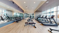 Located at 2132 City Gate Lane in Naperville, IL, this well-appointed fitness center offers a comprehensive and thoughtfully designed workout environment on a single level. The space is characterized by large, floor-to-ceiling windows along its perimeter that flood the interior with natural light and provide open views of the surrounding streetscape, creating an energizing atmosphere for exercise.

The light wood flooring throughout gives the space a clean, modern aesthetic, complemented by a slatted wood ceiling detail and recessed lighting that adds warmth and visual depth. Full-length mirrors are strategically positioned to expand the sense of space and allow users to monitor their form during workouts.

The cardio section is generously equipped with multiple treadmills, elliptical trainers, a rowing machine, and stationary bikes, all positioned to take advantage of the expansive street-facing windows. The strength training area features a comprehensive dumbbell rack with a wide range of weights, adjustable benches, and cable machines, offering users everything needed for a full resistance workout.

Accent walls in muted tones of blue-gray and taupe provide subtle visual contrast, while the open floor plan ensures ample space between equipment for comfortable movement. The overall ambiance is modern, clean, and welcoming, reflecting a premium facility designed to support a wide range of fitness goals.