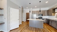 This well-appointed single-floor residence showcases a thoughtfully designed open-concept layout where the kitchen serves as a central focal point. The kitchen is outfitted with taupe-toned shaker-style cabinetry, light granite countertops, a full suite of stainless steel appliances including a refrigerator, electric range, and built-in microwave, along with a white tile backsplash. A central island with matching granite provides additional prep space and casual seating, illuminated by a pair of sleek black pendant lights. Warm hardwood-style flooring flows throughout the kitchen and main living areas, contrasting with plush carpet in the bedroom.

The living area is bright and airy, featuring recessed lighting, crown molding details, and a sliding glass door that draws in natural light and outdoor views. Built-in shelving adds a functional architectural detail. The bedroom benefits from large windows that invite abundant natural light. The bathroom continues the cohesive design language with matching cabinetry, granite countertops, a framed mirror, and hardwood-style flooring. Throughout the space, neutral gray walls and consistent finishes create a clean, modern aesthetic.