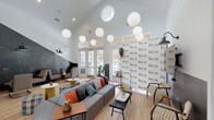 This dynamic single-floor community space for Bryn is designed to foster connection and comfort through thoughtful, eclectic design. The main lounge is the heart of the space, boasting soaring vaulted ceilings that create an airy, open atmosphere. A cluster of oversized globe pendant lights hangs dramatically overhead, casting a warm, diffused glow across the light hardwood floors below. Tall French doors and transom windows line the perimeter, flooding the interior with abundant natural light and offering views of the outdoors. The lounge is anchored by a generously sized curved sectional sofa accented with colorful throw pillows, complemented by plaid poufs and a round white coffee table — creating an inviting social hub. A signature branded accent wall featuring the "Bryn" logo adds a bold, graphic identity to the space. A striking diagonal chalkboard wall serves as both an artistic feature and an interactive community board, surrounded by retro-style cantilever armchairs and side tables for casual seating. Adjacent to the main lounge, a dedicated business center offers a long communal work surface with multiple computer stations, teal-painted walls, stylish pendant lighting, and carpeted flooring — providing a focused, professional environment within the broader community setting.
