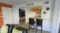 This single-floor residence offers an open and functional layout that connects the main living spaces in a cohesive flow. The living area is carpeted and furnished with a dark sofa accented by light-colored cushions, creating a casual and comfortable atmosphere. A peninsula-style breakfast bar with a light granite-look countertop serves as the dividing element between the living space and kitchen, accompanied by two dark leather-upholstered bar stools that provide convenient seating.

The kitchen, positioned in a recessed nook, features warm honey-toned cabinetry, a white refrigerator, and a range. Track lighting illuminates the workspace efficiently. The design of the pass-through opening between the kitchen and living area enhances the sense of openness throughout the space.

A doorway visible from the main living area leads directly into the bedroom, which is carpeted and includes a bed with neutral bedding. The room appears to receive ambient light, contributing to a relaxed feel. Central air supply vents are visible in the ceiling, indicating a centralized HVAC system throughout. Decorative wall accents add a finishing touch to the overall aesthetic.