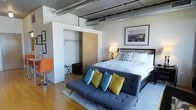 This urban loft-style studio showcases a thoughtfully designed open-concept layout that blends industrial character with modern comfort. The most striking architectural feature is the exposed silver ductwork running along the ceiling, lending the space a distinctly industrial aesthetic that is both functional and visually dynamic.

Warm hardwood flooring flows throughout, grounding the space and complementing the clean white walls. The sleeping area is anchored by a generously sized bed dressed in crisp white linens, positioned beneath a framed black-and-white cityscape print that adds a classic urban touch. A cozy gray futon sofa, adorned with vibrant teal and yellow accent pillows, sits at the foot of the bed, effectively delineating the living and sleeping zones within the open floor plan.

A built-in closet enclosure with sliding wood-panel doors provides smart, integrated storage, maintaining the clean lines of the interior design. Soft ambient lighting from a bedside lamp creates a warm, inviting atmosphere throughout the space.

Overall, this studio offers an efficient yet stylish living environment, balancing industrial design elements with warm, residential touches to create a space that feels both contemporary and comfortable.