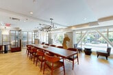Located at 1440 Beacon Street in Brookline, MA, this elegantly designed single-floor amenity space offers a refined blend of functionality and modern aesthetics. The main gathering area centers around a substantial hardwood conference table surrounded by mid-century modern chairs, illuminated by a striking sculptural chandelier with globe fixtures. Light-toned herringbone hardwood flooring runs throughout, lending warmth and visual continuity across the space.

Floor-to-ceiling windows along the street-facing wall bathe the interior in abundant natural light while offering views of the tree-lined boulevard outside. A curated selection of original artwork adorns the walls, contributing to the space's gallery-like ambiance. Decorative sculptural pieces are thoughtfully placed throughout, adding artistic character.

A semi-private lounge area with accent chairs and a low bowl table provides a relaxed seating alternative, while gold-framed frosted glass partitions define intimate booth-style seating areas equipped with built-in storage. A fully appointed kitchenette features marble countertops, white cabinetry, a stainless steel sink, and a Bosch appliance suite. An adjacent room accessible via glass-paneled doors offers additional private space. Recessed lighting and clean white ceilings complete the polished, contemporary atmosphere throughout.