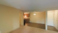 This single-floor space features an open layout that connects the living area to the kitchen, creating a cohesive and functional floor plan. The living area is finished with neutral-toned carpet, offering a comfortable and understated foundation that complements the warm beige walls. A textured ceiling runs throughout, adding a subtle dimensional quality to the interior.

The kitchen is positioned adjacent to the living space and is defined by medium-toned wooden cabinetry that provides ample storage. A countertop with a darker finish runs along the workspace, which includes a sink positioned beneath the upper cabinets. A pendant light fixture with a bowl-shaped shade hangs above, casting warm, focused light over the area and contributing to the overall warmth of the space. A dark-colored appliance, likely a refrigerator, is visible near the kitchen entry.

The transition between the carpeted living area and the hard flooring of the kitchen creates a clear delineation between the two zones while maintaining an open, connected feel. The neutral color palette and warm lighting throughout give the space a welcoming and practical ambiance suited for everyday living.