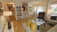 This single-floor residence offers a thoughtfully designed open-concept layout that seamlessly connects the main living areas. Rich hardwood flooring flows throughout, tying the space together with warmth and continuity. The living room serves as the heart of the home, featuring eclectic furnishings including a sleek coffee table, bold yellow accent stools, and a cushioned armchair, all anchored by a patterned area rug that adds texture and character. A well-stocked bookshelf acts as a natural divider between the living area and the kitchen, adding both function and charm. The kitchen is equipped with a raised breakfast bar and metal bar stools, ideal for casual dining or entertaining. Track lighting overhead ensures the kitchen remains bright and functional. Adjacent to the living room, a dedicated dining area benefits from abundant natural light streaming through large windows dressed with soft roman shades, centering around a round table with seating for several guests. Colorful artwork adorns the walls, contributing to the space's vibrant and inviting ambiance. The overall atmosphere is warm, eclectic, and well-appointed, offering a comfortable and stylish environment suited for both everyday living and entertaining.