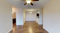 This single-floor residence offers a thoughtfully designed open-concept layout that seamlessly connects the living, dining, and kitchen areas. The main living space is finished with rich, dark hardwood-style flooring that runs throughout, lending a warm and grounded aesthetic to the interior. A ceiling fan with a light fixture anchors the living area, providing both comfort and ambient lighting, while a decorative chandelier marks the dining zone and adds an elegant touch to everyday living.

The open floor plan allows for generous sightlines across the space, creating a sense of openness and ease of movement. A pass-through window connects the main living area to the kitchen, which features dark wood cabinetry and what appears to be a stone or tile countertop — a functional and stylish detail that enhances the overall design. The kitchen cabinetry provides ample storage while maintaining a cohesive, refined look.

Neutral, light-toned walls throughout keep the space feeling bright and versatile, ready to accommodate a variety of interior styles. A glimpse into an adjacent room reveals additional dark cabinetry, suggesting a well-appointed bathroom or laundry area nearby. Floor vents, recessed lighting, and clean baseboards reflect attention to practical detail, making this a well-rounded and move-in-ready living environment.