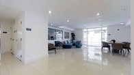 This single-floor space features a spacious, open-plan lounge area designed with a clean, contemporary aesthetic. The interior is characterized by large-format polished white tile flooring that reflects natural light throughout, contributing to an airy and expansive atmosphere. Floor-to-ceiling windows and glass panels line one side of the space, allowing abundant natural light to flood the interior while offering views of an outdoor balcony or terrace area beyond.

The lounge area is furnished with a modular blue sectional sofa arrangement, complemented by a round blue coffee table, and accented by a pair of traditional-style wooden armchairs that add a subtle contrast in texture and tone. A tall indoor plant provides a natural focal point near the seating area, adding warmth to the otherwise crisp, white interior.

Recessed ceiling lighting is evenly distributed across the flat white ceiling, ensuring well-balanced illumination throughout the space. The overall ambiance is bright, open, and polished, reflecting a modern design sensibility that emphasizes clean lines, neutral tones, and an uncluttered layout well-suited for a communal or reception-style environment.
