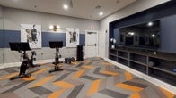 This single-level fitness studio is thoughtfully designed to support a wide range of workout styles. The space immediately draws attention with its striking chevron-patterned carpet tiles in orange, gray, and charcoal — a dynamic flooring choice that energizes the room and establishes a modern aesthetic throughout.

The walls are finished in a two-tone scheme, pairing a deep navy blue upper half with a lighter gray lower section, divided by crisp white chair rail molding. This color palette carries throughout the space, lending it a cohesive and professional appearance. Abstract wall art adds a creative accent without overwhelming the functional focus of the room.

A prominent built-in media niche, set within a navy-painted alcove, houses a large flat-screen television and open shelving beneath, providing both entertainment and storage. Full-length mirrors along one wall expand the sense of space and allow users to monitor their form during workouts.

Equipment is well-curated and includes a spin bike with an integrated display, kettlebells, a dumbbell rack, medicine balls and stability balls on a multi-tier rack, foam rollers, a ballet barre, and a plyo box. Recessed lighting illuminates the space evenly, and a glass entry door adds a sleek, modern touch to the overall design.