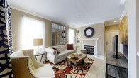 This single-floor living space offers a thoughtfully designed and inviting interior centered around a classic white-mantled fireplace, which serves as the room's focal point. The neutral, warm beige walls create a timeless backdrop that complements the carefully chosen furnishings, including a tufted cream sofa accented with decorative pillows and coordinating armchairs. Crown molding along the ceiling adds an architectural detail that elevates the overall aesthetic.

Natural light flows generously into the space through multiple windows dressed with sheer white curtains, contributing to the bright and airy atmosphere. A large multi-pane decorative mirror mounted on one wall further amplifies the sense of light and openness. Built-in shelving beside the fireplace provides functional display and storage space.

The room is grounded by a bold, patterned area rug in rich tones of red, brown, and cream, adding warmth and visual interest underfoot. A rustic wooden coffee table anchors the seating arrangement, completing the balanced mix of classic and casual elements. Overall, the space exudes a comfortable, well-appointed ambiance suitable for both relaxation and entertaining.