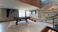 This single-floor amenity lounge is a masterclass in industrial-chic design, seamlessly blending raw materials with refined finishes to create an inviting and dynamic gathering space. Throughout, polished concrete floors provide a clean, durable foundation that complements the space's rugged character.

The main lounge area commands attention with soaring double-height ceilings, dramatic exposed Edison globe pendant lighting, and a striking feature wall clad in reclaimed wood planks. A vintage-style shuffleboard table anchors the room, surrounded by leather bar stools and accented by a bold, hand-illustrated chalkboard mural wall that adds artistic personality. Natural light filters in from clerestory windows above, enhancing the open, loft-like atmosphere.

An adjacent game room offers a dedicated pool table set against walls fully sheathed in reclaimed wood, with a wall-mounted flat-screen TV, a dart board, and generous natural light streaming through large windows with blinds — making it a comfortable retreat for recreation.

The wet bar and kitchenette area is thoughtfully appointed with dark cabinetry, marble-look countertops, a hexagonal mosaic tile backsplash, a stainless steel sink, and industrial pipe open shelving. A reclaimed wood accent wall adds warmth, while pendant lighting provides a focused, functional glow. Together, these spaces form a cohesive, well-designed entertainment environment with strong visual character.