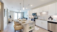 This single-story residence showcases a thoughtfully designed open-concept layout with clean, contemporary finishes throughout. The kitchen serves as a central hub, featuring crisp white cabinetry, a subway tile backsplash, stainless steel appliances, and a granite-topped island with bar seating illuminated by pendant lighting. Light wood-style flooring flows seamlessly into the living area, where large sliding glass doors invite natural light and provide outdoor access. A ceiling fan overhead and neutral wall tones contribute to a relaxed, airy atmosphere.

The bedroom is a well-appointed retreat, distinguished by a bold blue accent wall and natural light streaming through large windows with patterned drapery. A ceiling fan adds comfort, and direct access to the en-suite bathroom enhances convenience. The bathroom offers dual granite-topped vanities with white cabinetry, wood-style flooring, a large mirror with lighted surround, and a combined tub and tiled shower. Built-in open shelving provides additional storage. A spacious walk-in closet with multiple hanging rods and shelving completes the private quarters, offering generous and well-organized storage space.