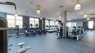 Located at The Vanderbilt in Westbury, New York, this fitness center offers a well-appointed and thoughtfully designed workout space. The gym is finished with durable rubber flooring in a cool slate-gray tone, providing a safe and comfortable surface for all types of exercise. A row of large windows lines the walls, flooding the space with an abundance of natural light and creating an open, energizing atmosphere. Elegant pendant lights with warm-toned shades hang from the ceiling, complementing the gym's refined aesthetic while ensuring consistent illumination throughout. The color palette of soft gray walls and neutral accents gives the space a clean, modern look. The fitness center is comprehensively equipped with a variety of cardio machines, including treadmills and elliptical cross-trainers, arranged to take full advantage of the natural light from the windows. A dedicated strength training area features multi-use cable and resistance machines, along with free weights, catering to a wide range of workout routines. Full-length mirrors are integrated along one wall, enhancing the sense of space and allowing members to monitor their form. The overall ambiance is one of a well-maintained, upscale fitness facility designed to support a complete and effective exercise experience.