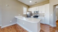 Located at 12821 Little Penny Dr in Huntersville, NC, this single-floor 2-bedroom, 2-bathroom home offers a thoughtfully designed layout with a clean, modern aesthetic throughout. The open-concept kitchen and living area serves as the heart of the home, showcasing warm hardwood-style flooring, white shaker cabinetry, a mosaic tile backsplash, and a large peninsula with a white countertop and undermount sink — ideal for both cooking and casual dining. Stainless steel appliances complete the kitchen's contemporary appeal. The living room is a standout, featuring soaring ceilings and a pair of oversized arched windows that flood the space with natural light and offer views of the surrounding neighborhood. Both bedrooms are carpeted for comfort, with the primary bedroom offering generous proportions and ample natural light. The two bathrooms are finished with wood-look flooring, white vanities, and full tub-shower combinations with tile surrounds. Neutral, warm-toned walls throughout create a cohesive and welcoming ambiance, while recessed lighting and ceiling fans add functional comfort. Two hallways connect the spaces efficiently, and a dedicated closet provides additional storage.