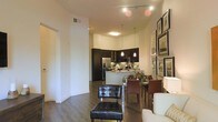 This single-floor residence showcases an open-concept layout that seamlessly connects the living, dining, and kitchen areas into one cohesive, functional space. The kitchen is outfitted with dark espresso cabinetry that contrasts sharply against light countertops, stainless steel appliances including a French door refrigerator, and a breakfast bar that doubles as a casual dining surface. Pendant lighting hangs above the kitchen area, adding focused illumination and a stylish touch. The dining area accommodates a full table with leather seating, positioned adjacent to the kitchen for easy flow during meals. Track lighting on the ceiling provides adjustable illumination across the open space, complementing the recessed lighting throughout. Dark hardwood flooring runs continuously through the main areas, grounding the space with warmth and visual cohesion. A tufted leather accent chair anchors the living area, suggesting a comfortable and well-appointed lounge space. A gallery wall featuring framed artwork adds a curated, polished aesthetic to one side of the room. The overall ambiance is modern and sophisticated, with a neutral color palette highlighted by dark accent tones.