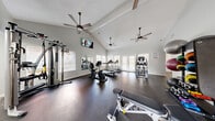 This single-story fitness center offers a thoughtfully designed workout environment across a compact yet efficiently organized footprint. The space is divided into two areas: a main exercise room and a connecting entryway/transition corridor with light wood-look flooring.

The primary workout area is distinguished by soaring vaulted ceilings that create an open, airy atmosphere, complemented by multiple ceiling fans that promote air circulation throughout the space. Dark rubber flooring covers the exercise area, providing both durability and safety. Large windows fitted with blinds line the walls, while a set of glass French doors at the far end draws in abundant natural light and offers views to the outdoors.

The facility is well-stocked with a comprehensive range of equipment, including True-brand treadmills, a recumbent bike, a stair climber, and a multi-station cable machine. A dedicated free weights section features a full dumbbell rack alongside a weight bench. A medicine ball and kettlebell storage rack rounds out the functional training options. Wall-mounted mirrors expand the perceived space visually, and mounted flat-screen televisions provide entertainment. The neutral white walls and clean design maintain a bright, motivating environment.