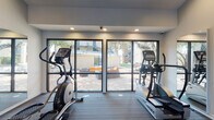 Located in Austin, TX, this community offers a carefully curated combination of indoor fitness amenities and inviting outdoor living spaces. The fitness center is bright and functional, featuring large floor-to-ceiling windows framed in dark metal that flood the room with natural light while providing views of the outdoor courtyard. Cardio equipment, including ellipticals and stair climbers, is positioned to take advantage of these views, creating an energizing workout environment with wood-style flooring and recessed lighting overhead.

The outdoor grounds are a standout feature, designed with both aesthetics and relaxation in mind. A central lawn area is accented by a linear water feature with smooth river rocks, creating a tranquil focal point. Concrete stepping stone pathways guide residents through well-maintained turf, ornamental grasses, and layered plantings. A pergola draped in lush climbing vines adds natural shade and architectural character. Adirondack-style seating is thoughtfully placed throughout the patio areas, offering comfortable spots to unwind beneath the canopy of mature oak trees that are characteristic of Austin's natural landscape.