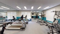 Located at 2400 Garden Hill Dr in Raleigh, NC, this dedicated fitness center offers a comprehensive workout environment designed to accommodate a wide range of exercise routines. The space is laid out on a single floor and is organized to maximize functionality and flow between equipment zones.

The cardio area features multiple True-brand treadmills arranged for easy access, complemented by additional aerobic machines including a stationary bike and elliptical-style equipment. Full-length mirrors line one wall, visually expanding the space and allowing users to monitor their form during exercise. Recessed fluorescent panel lighting illuminates the entire area evenly, while blinds-fitted windows along the perimeter invite natural light and provide ventilation.

The strength training section is well-appointed with a multi-function cable and weight machine, a power tower for bodyweight exercises, a flat workout bench, and a rack of dumbbells spanning a variety of weights. A Paramount multi-station gym unit rounds out the resistance training offerings.

A dedicated functional training corner holds medicine balls, stability balls in assorted sizes, resistance bands, and additional accessories displayed on wall-mounted storage. A wall-mounted flat-screen television is positioned for viewing during workouts. The two-tone wall design — neutral gray above a bold teal accent band — gives the space a clean, energetic aesthetic consistent with a professional fitness facility.