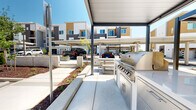 Located in Folsom, CA, this well-appointed outdoor amenity area serves as a central gathering space for the surrounding residential community. The space is thoughtfully designed with a combination of covered and open-air zones, providing flexibility for both casual relaxation and social entertaining throughout the year.

At the heart of the area is a sleek, covered outdoor kitchen station equipped with a stainless steel built-in grill and cabinetry, offering a fully functional setup for barbecuing. Adjacent to the grilling zone, multiple pergola structures with clean-lined metal frames provide shaded seating areas furnished with tables and chairs, ideal for outdoor dining and conversation.

Concrete hardscaping forms broad, level walkways that connect the various activity zones, while raised planter beds filled with mulch and young ornamental trees and shrubs introduce greenery and define distinct spaces throughout the courtyard. A dedicated fire pit area adds another dimension for evening gatherings.

The surrounding architecture features modern multi-story residential buildings with a contemporary palette of white, gray, and warm tan tones, creating a cohesive and attractive backdrop. Covered parking canopies are integrated nearby, adding practical convenience. The overall ambiance is clean, modern, and community-oriented, designed to encourage outdoor living under the abundant California sunshine.