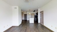 This single-floor unit presents an open-concept living and kitchen layout designed for functional everyday living. The space is finished with rich, dark hardwood-style flooring that extends throughout, providing a warm contrast against the clean white walls and ceilings. The kitchen is positioned toward the center-rear of the main living area and features dark cabinetry, stainless steel appliances including a refrigerator and range, and a white kitchen island that doubles as a casual dining or prep surface. A pendant light fixture hangs above the island, adding a decorative focal point to the cooking area. Track lighting is also visible overhead, offering flexible illumination across the space. The open layout allows the living area to flow naturally toward the kitchen, creating a connected and sociable atmosphere. A doorway near the kitchen leads to an additional room, while a secondary door is visible at the far end of the unit, likely serving as the main entry. The overall design aesthetic is clean and contemporary, with neutral tones and modern finishes throughout.