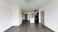 This single-floor unit presents an open-concept living and kitchen layout designed for functional everyday living. The space is finished with rich, dark hardwood-style flooring that extends throughout, providing a warm contrast against the clean white walls and ceilings. The kitchen is positioned toward the center-rear of the main living area and features dark cabinetry, stainless steel appliances including a refrigerator and range, and a white kitchen island that doubles as a casual dining or prep surface. A pendant light fixture hangs above the island, adding a decorative focal point to the cooking area. Track lighting is also visible overhead, offering flexible illumination across the space. The open layout allows the living area to flow naturally toward the kitchen, creating a connected and sociable atmosphere. A doorway near the kitchen leads to an additional room, while a secondary door is visible at the far end of the unit, likely serving as the main entry. The overall design aesthetic is clean and contemporary, with neutral tones and modern finishes throughout.