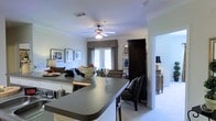 This single-floor space showcases an open-concept design that seamlessly connects the kitchen and living area, creating a cohesive and functional layout. The kitchen features a dark-toned countertop peninsula that serves as both a workspace and a casual dining or entertaining surface, complemented by light cabinetry and modern fixtures. Crown molding runs throughout the space, adding a refined architectural detail to the otherwise clean, white walls and ceilings. A ceiling fan with a built-in light fixture is centrally positioned above the living area, providing both comfort and illumination. The living space is carpeted and furnished with dark wood pieces, including a prominent armoire and accent seating, contributing to a warm, traditional aesthetic. Drapes with decorative valances frame a set of glass doors that allow natural light to filter into the space and offer a view to the outdoors. An adjacent room, visible through a doorway, hints at additional living or bedroom space, furnished with a dresser and lamp. Overall, the space carries a classic, well-appointed ambiance with thoughtful details throughout.