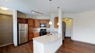 This single-floor residence offers a thoughtfully designed layout centered around a stylish, open-concept kitchen. The kitchen is a standout feature, showcasing warm medium-toned wood cabinetry that provides ample storage, paired with sleek granite countertops in soft neutral tones. A curved kitchen island serves as both a functional workspace and a natural gathering point, complete with an undermount sink and elegant pendant lighting above. Black appliances, including a range and built-in microwave, contrast beautifully with the stainless steel refrigerator, adding a contemporary touch. Track lighting on the ceiling further enhances the kitchen's bright and functional ambiance. Rich hardwood flooring runs throughout the space, adding warmth and visual continuity. An arched doorway visible in the background adds an architectural detail that lends character and a sense of openness to the floor plan, guiding the eye through the space. The clean white walls keep the interior feeling light and airy, while the overall layout promotes a smooth and intuitive flow between living areas. This residence combines practical design with refined finishes to create a comfortable and inviting living environment.