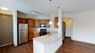 This single-floor residence offers a thoughtfully designed layout centered around a stylish, open-concept kitchen. The kitchen is a standout feature, showcasing warm medium-toned wood cabinetry that provides ample storage, paired with sleek granite countertops in soft neutral tones. A curved kitchen island serves as both a functional workspace and a natural gathering point, complete with an undermount sink and elegant pendant lighting above. Black appliances, including a range and built-in microwave, contrast beautifully with the stainless steel refrigerator, adding a contemporary touch. Track lighting on the ceiling further enhances the kitchen's bright and functional ambiance. Rich hardwood flooring runs throughout the space, adding warmth and visual continuity. An arched doorway visible in the background adds an architectural detail that lends character and a sense of openness to the floor plan, guiding the eye through the space. The clean white walls keep the interior feeling light and airy, while the overall layout promotes a smooth and intuitive flow between living areas. This residence combines practical design with refined finishes to create a comfortable and inviting living environment.
