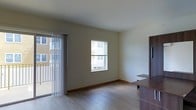 This single-floor unit offers a clean, functional layout designed with modern, understated finishes throughout. The main living area is highlighted by smooth wood-style flooring that runs throughout the space, contributing to a warm and cohesive aesthetic. Natural light flows in through a double-hung window positioned along the exterior wall, while vertical blinds frame a sliding glass door that opens to a private balcony — offering a connection to the outdoors and views of the surrounding building exterior. The walls are finished in a neutral, light tone that enhances the brightness of the room and provides a versatile backdrop for furnishings. A dark wood-toned built-in or furniture piece is visible along one wall, adding a contrasting design element to the otherwise light interior. The space maintains a minimalist character, with clean lines and an open feel that allows for flexible furniture arrangements. The combination of the balcony access, ample natural light, and practical flooring makes this a well-appointed and functional living space suited for everyday comfort.