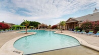 Located at 200 Wickerleaf Way in Garner, NC, this community amenity space offers a resort-inspired outdoor experience centered around a large, freeform swimming pool with a curvilinear design. The pool area is generously appointed with rows of lounge chairs, vibrant canopied cabanas in green and blue, and lush tropical palm trees that contribute to a relaxed, vacation-like atmosphere. Colorful crape myrtle trees and manicured landscaping frame the perimeter, adding seasonal color and visual appeal.

Adjacent to the pool, a brick-constructed clubhouse building anchors the space architecturally, featuring a traditional pitched roofline and classic exterior detailing. A screened-in porch extends from the clubhouse, furnished with wicker dining and lounge seating, ceiling fans, and floor-to-ceiling screened windows that provide panoramic views of the pool and surrounding grounds — ideal for outdoor gatherings in any weather.

The interior portion of the amenity building includes a kitchen-style area with dark cabinetry, rich granite countertops, stainless steel appliances, and tall ceilings with clean white walls. An overhead electrical panel is also accessible within this space. Together, these features make this a well-equipped and visually appealing community hub designed for both relaxation and social gatherings.