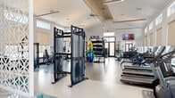 This spacious, single-floor fitness center offers a thoroughly modern workout environment designed to accommodate a wide variety of exercise routines. The facility's architecture is defined by soaring ceilings with decorative tray detailing, large clerestory windows lining the upper walls, and an open floor plan that creates an airy, energizing atmosphere throughout. Dual ceiling fans enhance comfort across the workout floor, which features durable, speckled epoxy flooring throughout.

The cardio section is well-appointed with treadmills, ellipticals, stair climbers, and indoor cycling bikes, many equipped with integrated digital displays. A dedicated strength training area houses a full rack of dumbbells, weight benches, and a selection of Matrix selectorized machines targeting various muscle groups. Functional training is thoroughly supported with a multi-purpose rig, TRX suspension trainers, kettlebells, battle ropes, medicine balls, resistance bands, and yoga mats stored on a colorful organizational tower.

Cable machines, including a dual-adjustable pulley system, anchor the center of the space, while large wall mirrors amplify the sense of openness. Natural light pours in through expansive windows offering views of the surrounding outdoor area. A decorative lattice accent wall and wall-mounted televisions add visual interest. The entrance features glass double doors, completing a bright, welcoming, and fully equipped fitness facility.