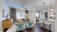 This single-story residence is thoughtfully designed with an open-concept layout that seamlessly connects the living, dining, and kitchen areas. Rich dark hardwood flooring flows throughout the main living spaces, anchored by a decorative column that subtly defines zones without interrupting the open feel. The living room features a ceiling fan, multiple windows with blinds and draping curtains, and ample natural light. Two light blue accent chairs and a neutral sofa create a welcoming seating arrangement atop a geometric area rug.

The kitchen is outfitted with white shaker-style cabinetry, granite countertops, a mosaic tile backsplash, stainless steel appliances including a range, microwave, and refrigerator, and a peninsula with pendant lighting overhead. A dining area sits adjacent, completing the open entertaining space.

Both bedrooms are carpeted and generously sized, each with ceiling fans, large windows, and walk-in closets. The primary bedroom connects to a bathroom featuring granite vanity counters, white cabinetry, dark hardware, and a tub-shower combination. The second bathroom mirrors this design with similar finishes and its own walk-in closet access, offering a cohesive and polished aesthetic throughout.