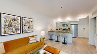 This thoughtfully designed 1-bedroom, 1-bathroom unit offers an efficient and modern layout across a single floor. The open-concept living and kitchen area serves as the heart of the unit, where light-toned hardwood flooring flows seamlessly throughout. The kitchen is equipped with stainless steel appliances, including a refrigerator, electric range, and over-the-range microwave, complemented by light grey cabinetry, a tile backsplash, and a dark countertop peninsula. Three white pendant lights with gold interiors hang above the peninsula, which features bar seating for casual dining. The living area is bright and welcoming, anchored by large windows that frame views of lush greenery and an outdoor space, filling the room with natural light. The bedroom is a comfortable retreat, featuring a ceiling fan, large windows overlooking outdoor greenery, and ample room for a full-sized bed and furnishings. The bathroom offers a clean, contemporary design with a white vanity, dark countertop, a large mirror, and a full-sized shower-tub combination with white tile surround. A hallway connects the main living areas to the private spaces, completing the unit's functional and cohesive layout.