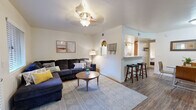 Located on E Prince Rd in Tucson, Arizona, this well-appointed 1-bedroom, 1-bathroom home offers an efficient and comfortable single-floor layout. The open-concept living room and kitchen area form the heart of the home, featuring durable hardwood-style flooring, a ceiling fan with light fixture, and natural light from multiple windows. The living room provides a welcoming atmosphere with ample space for seating and entertainment, including a wall-mounted TV area and a front entry door with adjacent window. A charming arched passageway connects the living area to the kitchen, which is outfitted with warm wood cabinetry, marble-style countertops, a full suite of white appliances including a refrigerator, electric range, and built-in microwave, and a breakfast bar with counter seating. The spacious bedroom features plush carpeting, a ceiling fan, a generous closet, and a window that allows natural light to filter in. The bathroom includes a full tub-shower combination with clean white tile surround. A hallway connects the main living spaces, completing this functional and thoughtfully arranged Tucson home.