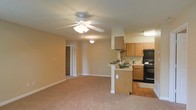 This single-floor residence features an open-concept layout that seamlessly connects the living area and kitchen, creating a spacious and functional environment. The living area is finished with plush wall-to-wall carpeting and warm beige walls, contributing to a welcoming ambiance. A ceiling fan with an integrated light fixture serves as a central feature, providing both ventilation and illumination throughout the space.

The kitchen is thoughtfully designed with medium-toned wooden cabinetry offering generous storage, and is equipped with black appliances including a range and oven with an overhead exhaust hood. A peninsula with a raised breakfast bar creates a natural division between the kitchen and living area while also providing additional counter space and casual seating. The kitchen area transitions to hardwood flooring, distinguishing it from the carpeted living zone.

A pendant light adds a decorative touch near the kitchen, while recessed lighting under the cabinetry ensures functional task lighting. Visible from the main living space is a hallway leading to a walk-in closet, adding notable storage capacity. The textured ceiling throughout adds subtle architectural character to the overall design.