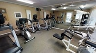 This fitness center offers a focused and functional workout environment housed within a single-floor facility. The space is outfitted with a comprehensive selection of cardio equipment, including multiple TRUE-brand elliptical trainers and treadmills arranged in an organized layout that maximizes the available floor space. A dedicated strength area features a weight bench and additional resistance equipment, catering to a well-rounded exercise routine. Large floor-to-ceiling mirrors line one wall, creating a sense of openness and allowing users to monitor their form during workouts. The flooring consists of durable, cushioned rubber material, providing both safety and comfort underfoot. Overhead fluorescent lighting ensures the space is bright and well-illuminated throughout. The walls are finished in a warm neutral tone, contributing to an inviting and energizing atmosphere. A wall-mounted speaker system is visible, suggesting the space is equipped with audio for an enhanced workout experience. Informational signage is posted near the entrance, offering guidance for facility use. Natural light filters in through a window, adding a refreshing element to the interior. Overall, the fitness center is clean, well-maintained, and thoughtfully arranged to accommodate a variety of exercise needs.