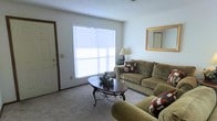 This single-floor residence offers a warm and inviting atmosphere, highlighted by a well-appointed living room that serves as the heart of the space. The living room features plush wall-to-wall carpeting in a neutral tone, creating a comfortable and cohesive foundation throughout. A large window dressed with horizontal blinds allows abundant natural light to fill the room, lending an open and airy feel. The space is furnished with a pair of generously sized, earth-toned sofas arranged around an elegant oval coffee table with ornate wrought-iron legs, making it ideal for relaxed gatherings. A gold-framed mirror adorns one wall, adding a classic decorative touch while amplifying the sense of space. A traditional floor lamp provides additional warm ambient lighting. The textured ceiling adds subtle architectural character to the room. A clearly defined entryway with a dark wood-trimmed door provides a natural transition into the living area, contributing to the overall sense of a well-organized and functional floor plan. The overall ambiance is cozy, classic, and move-in ready, with thoughtful design details that make the space feel both livable and welcoming.