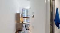 Located at 229 Duffield Street in the heart of Downtown Brooklyn, this private room offers a thoughtfully designed, efficient living space across a single floor. The main room serves as both a bedroom and functional living area, featuring warm hardwood flooring, bright white walls, and large floor-to-ceiling windows framed in rich wood tones that flood the space with natural light and provide views of the surrounding urban skyline. A built-in workstation with under-cabinet lighting, integrated shelving, and multiple drawers offers a dedicated area for work or study. Alongside the desk sits a compact stainless steel refrigerator and microwave unit, providing convenient in-room kitchen amenities. The sleeping area accommodates a full-sized bed and is positioned adjacent to the windows, enhancing the airy feel of the room. A dedicated closet with built-in hanging rods and shelving provides organized storage. The private bathroom is finished with neutral tile walls and flooring, and includes a glass-enclosed shower, toilet, and a wall-mounted vanity shelf. The overall design is clean and modern, making efficient use of every square foot while maintaining a comfortable and functional atmosphere suited for urban living.