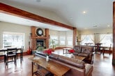 This well-appointed single-floor space offers a generous, open layout that seamlessly connects the living room, dining area, and kitchen into one cohesive environment. The interior is defined by its striking architectural character, most notably the bold, richly stained wood columns and ceiling beams that frame the open-plan living area and lend a rustic yet refined warmth throughout.

The living room features vaulted ceilings, a corner stone fireplace with a substantial wood mantel, and a sweeping bank of windows that floods the space with natural light and provides broad exterior views. The polished, stained concrete floors in deep amber and mahogany tones extend throughout, grounding the space with a durable and visually rich surface.

The dining area sits adjacent to the living room, benefiting from large windows dressed with sheer linen curtains that soften the natural light. A substantial wood dining table anchors this zone, making it well-suited for gatherings.

The kitchen is efficiently designed, featuring warm knotty wood cabinetry, dark countertops, stainless steel appliances, and a window above the sink that brings in additional daylight. The overall ambiance of the space is inviting and character-rich, blending natural materials, abundant light, and an open, connected floor plan into a highly functional and visually cohesive environment.